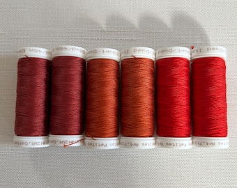 May include: Six spools of thread in shades of red and orange are arranged in a row. The thread is wound around white spools with printed text. The colors range from deep burgundy to bright red, suggesting a gradient.