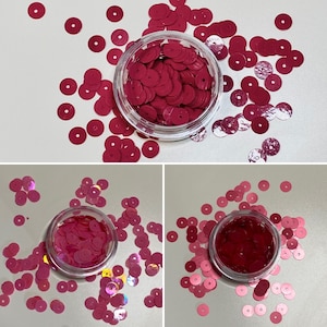 May include: A close-up image of a jar filled with small, pink, iridescent sequins. The sequins are scattered around the jar on a white surface.