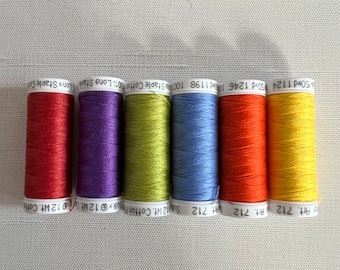 May include: Six spools of colorful sewing thread in red, purple, green, blue, orange, and yellow. Each spool is labeled with text including "100% Long Staple Cotton" and "Art. 712". The threads are arranged in a row.