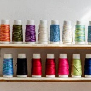 May include: A collection of spools of thread in various colours, including red, blue, green, white, and black. The spools are arranged in two rows on a wooden shelf.