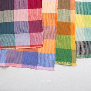 May include: Four overlapping fabric swatches featuring a woven checkered pattern in a vibrant array of colours including red, purple, teal, orange, yellow, green, and blue. The textile has a visible grid texture with frayed edges and the text FABLEISM.