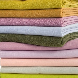 Può includere: Una pila di tessuti piegati in vari colori, tra cui giallo, oro, lavanda, azzurro, verde oliva e rosa. I tessuti sembrano essere di lino o un materiale simile strutturato, con motivi di tessitura visibili. La pila è ben disposta, mostrando le diverse tonalità.