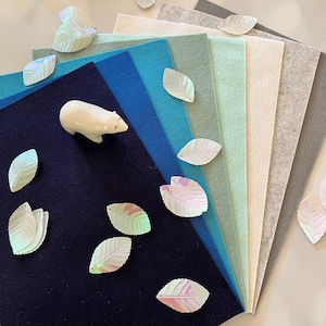 May include: A stack of felt fabric sheets in various shades of blue, green, and grey. The sheets are arranged in a fan shape with the darkest blue sheet at the bottom. There are iridescent white leaf-shaped sequins scattered around the sheets.