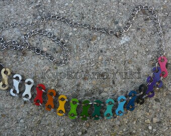 May include: A colorful necklace made from repurposed bicycle chain links. The links are painted in a rainbow pattern, with silver chain links connecting them. The necklace is lying on a gray concrete surface.