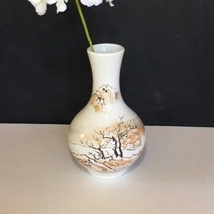 May include: A white ceramic vase with a landscape design featuring trees and a building, holding a white flower arrangement. The vase has a narrow neck and a rounded body, with the artwork in shades of brown and tan.