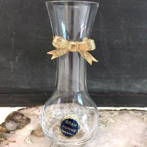 May include: A clear glass vase with a gold-colored bow around the neck. The vase has a bulbous base and a flared top. A blue and gold sticker on the base reads "GRAM FLORENCE MADE IN ITALY".