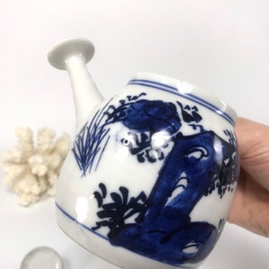 May include: A white ceramic teapot with a spout and handle, decorated with blue floral and landscape designs. The teapot has a small, round lid and is held in a hand. The teapot is approximately 4 inches tall.