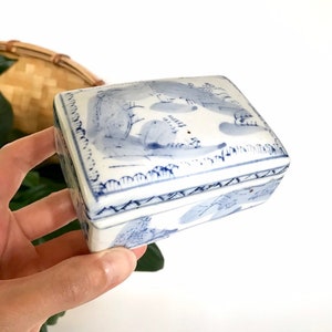 May include: A rectangular, white ceramic trinket box with a blue landscape design. The box has a lid and is held by a hand. The design features trees and other natural elements. The box is a decorative item.