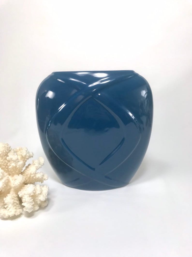 May include: A glossy, navy blue ceramic vase with a geometric, quilted pattern. The vase has a rounded shape and a wide opening. A piece of white coral is in the background.