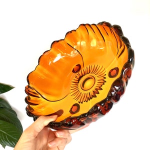 May include: Amber-colored glass bowl with a decorative sunburst design in the center. The bowl has a scalloped edge and is held against a white background. The bowl is a decorative piece.