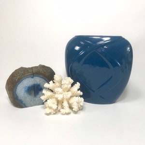 May include: A still life arrangement featuring a blue vase with a geometric design, a piece of white coral, and a blue and brown geode. The objects are arranged on a white surface.