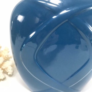 May include: A glossy, blue ceramic vase with a unique woven design. The vase has a rounded shape and a smooth, reflective surface. The color is a deep, muted blue, and the design creates a sense of depth and texture.