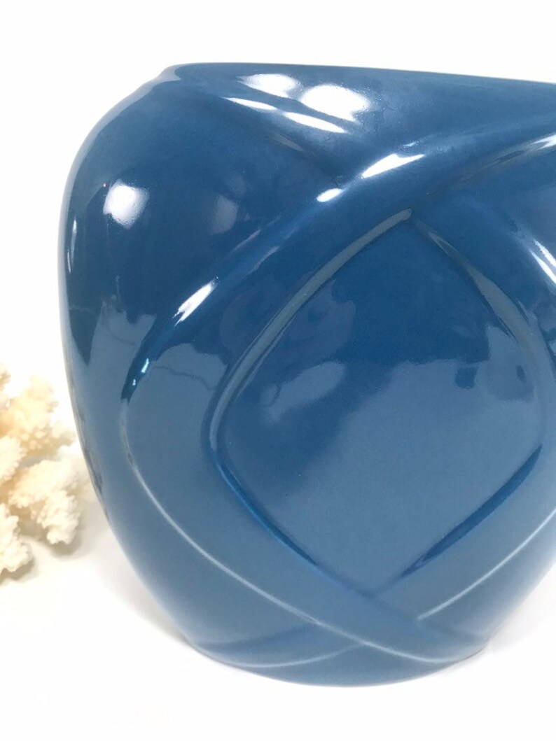 May include: A glossy, blue ceramic vase with a unique woven design. The vase has a rounded shape and a smooth, reflective surface. The color is a deep, muted blue, and the design creates a sense of depth and texture.