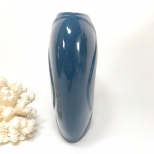 May include: A glossy, navy blue vase with a curved, elongated shape. The vase has a smooth, reflective surface and a slightly flared top. A piece of white coral is in the background.