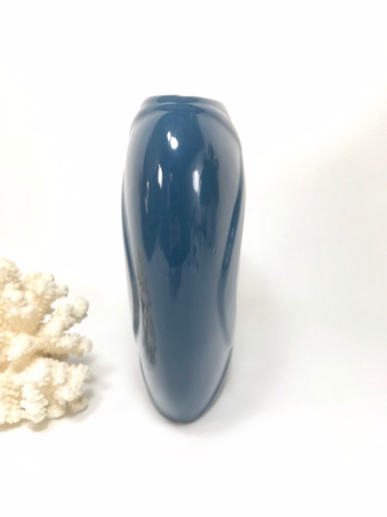 May include: A glossy, navy blue vase with a curved, elongated shape. The vase has a smooth, reflective surface and a slightly flared top. A piece of white coral is in the background.