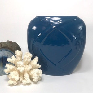 May include: A glossy, navy blue ceramic vase with a diamond pattern. A white coral and a piece of blue and gray stone are in the foreground. The vase has a wide, rounded shape and a smooth, reflective surface.