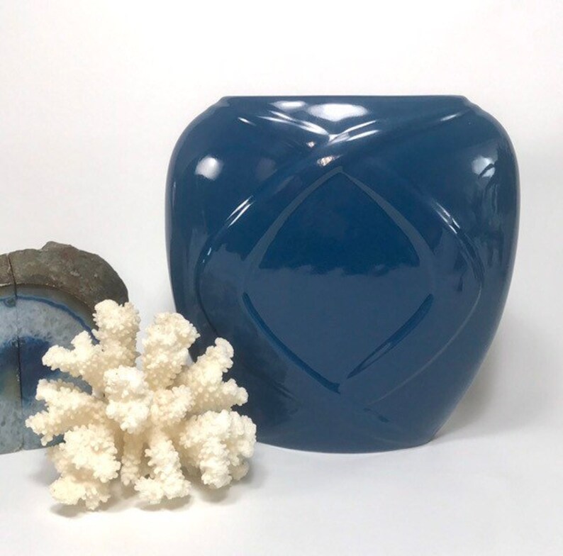 May include: A glossy, navy blue ceramic vase with a diamond pattern. A white coral and a piece of blue and gray stone are in the foreground. The vase has a wide, rounded shape and a smooth, reflective surface.