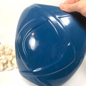 May include: A glossy, deep blue ceramic vase with a geometric design. The vase has a rounded shape with a series of intersecting lines creating a textured surface. The vase is held by a hand, and a piece of white coral is in the background.