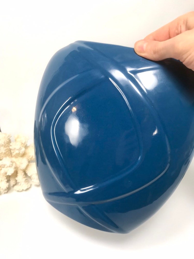 May include: A glossy, deep blue ceramic vase with a geometric design. The vase has a rounded shape with a series of intersecting lines creating a textured surface. The vase is held by a hand, and a piece of white coral is in the background.
