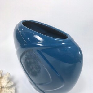 May include: A glossy, blue ceramic vase with a unique, asymmetrical shape. The vase has a wide, oval opening and a smooth, curved design. The color is a deep, solid blue, and the surface reflects light.