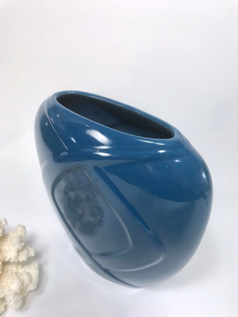 May include: A glossy, blue ceramic vase with a unique, asymmetrical shape. The vase has a wide, oval opening and a smooth, curved design. The color is a deep, solid blue, and the surface reflects light.