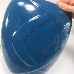 May include: A glossy, blue ceramic vase with a unique, curved design. The vase has a smooth, reflective surface and a modern aesthetic. The shape is reminiscent of a stylized helmet or a rounded, asymmetrical form. The vase is held by a hand.