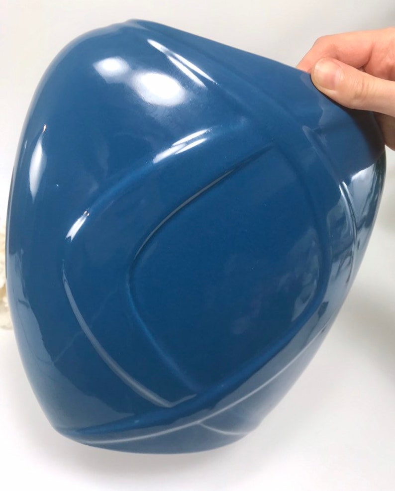 May include: A glossy, blue ceramic vase with a unique, curved design. The vase has a smooth, reflective surface and a modern aesthetic. The shape is reminiscent of a stylized helmet or a rounded, asymmetrical form. The vase is held by a hand.
