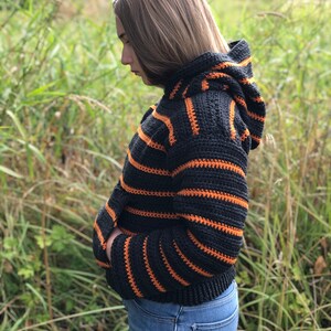 May include: A black and orange striped crocheted hooded sweater with long sleeves. The sweater is cropped and has a relaxed fit.