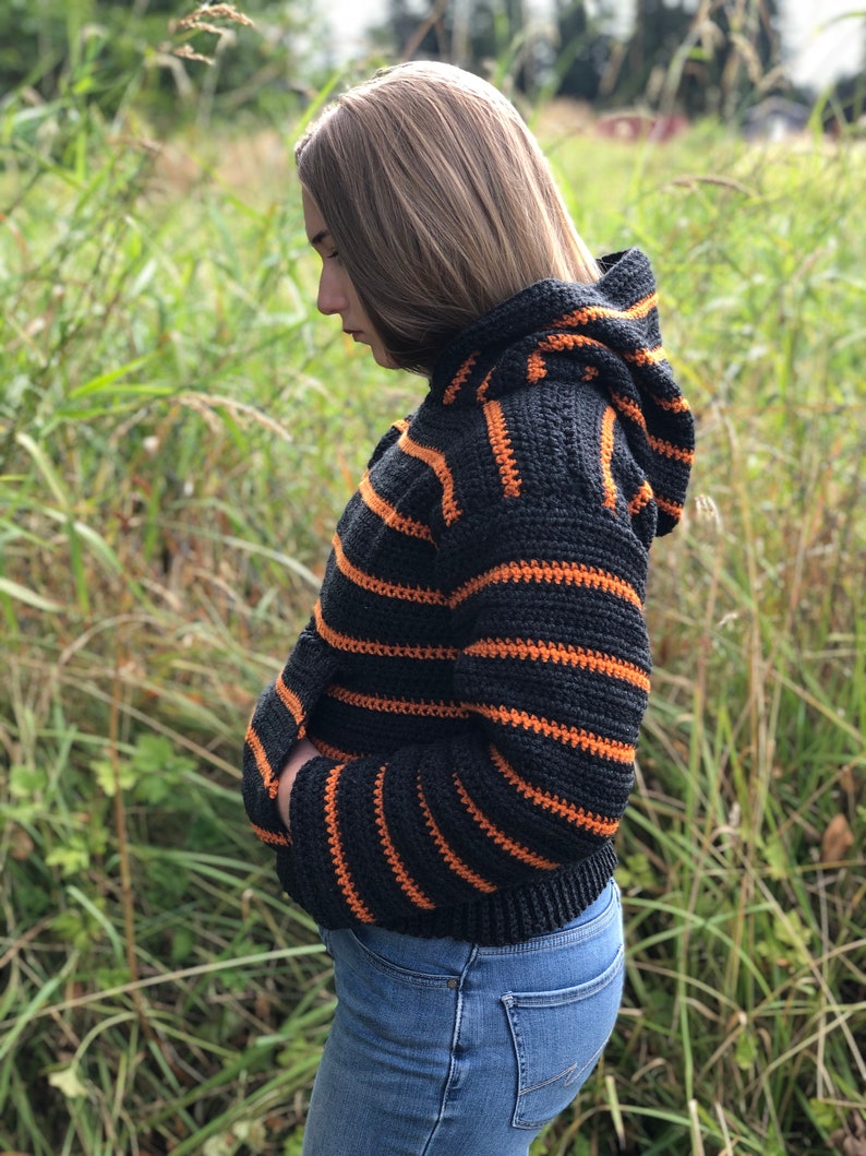 May include: A black and orange striped crocheted hooded sweater with long sleeves. The sweater is cropped and has a relaxed fit.