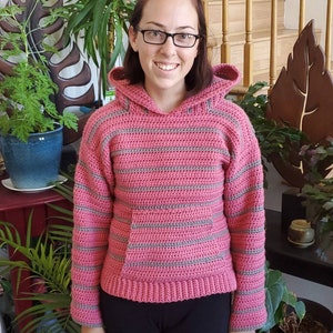 May include: A pink and gray striped crocheted hooded sweater with a pocket. The sweater is worn by a person with brown hair and glasses.