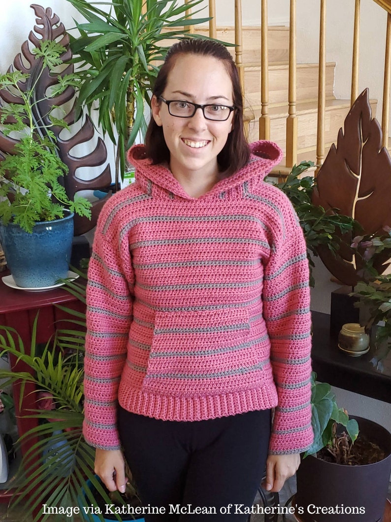 May include: A pink and gray striped crocheted hooded sweater with a pocket. The sweater is worn by a person with brown hair and glasses.