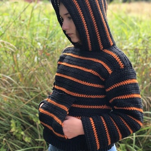 May include: A black and orange striped crocheted hoodie with a hood. The hoodie has a pocket on the front.