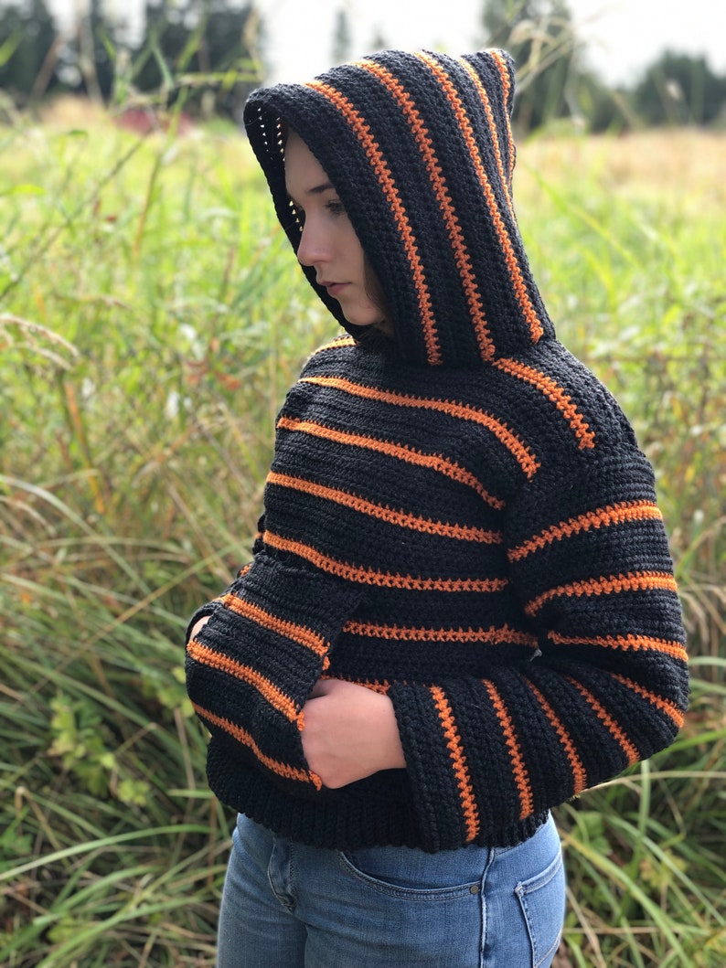 May include: A black and orange striped crocheted hoodie with a hood. The hoodie has a pocket on the front.