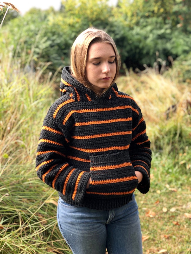 May include: A black and orange striped crocheted hoodie with a pocket. The hoodie has a hood and long sleeves.