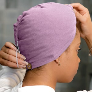 Pode incluir: Um turbante de cetim roxo com uma faixa elástica branca e um fecho de botão. O turbante está sendo usado por uma pessoa com cabelo escuro.