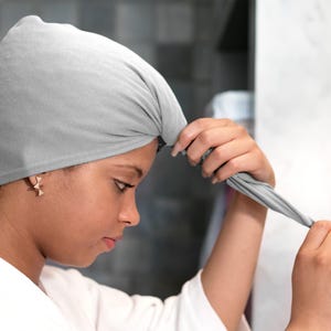 May include: A light grey hair towel is being secured on the head. The towel is made of a soft material and has a button closure. The person is wearing a white robe and gold earrings.