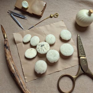 May include: A selection of handmade, cream-coloured fabric buttons on brown paper. The image also features gold scissors, a wooden-handled tool, and other sewing implements, indicating a craft or DIY project.