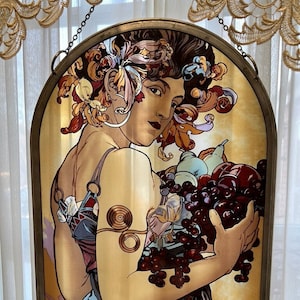 May include: A stained glass window depicting a woman with flowing hair and a floral crown, holding a basket of fruit. The woman is wearing a blue dress and the background is a warm yellow. The artist's signature, "Mucha", is visible in the bottom right corner.