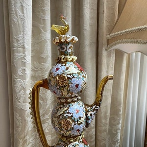 May include: A decorative glass teapot with a bird finial. The teapot is decorated with floral patterns in shades of blue, white, and gold on a brown background.