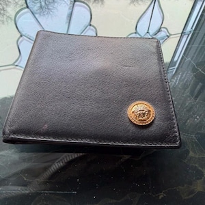 May include: Black leather wallet with a gold-colored emblem on the front. The wallet has a rectangular shape with a slightly angled top. The emblem appears to be a circular medallion with a detailed design.