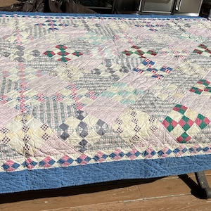May include: A vintage quilt with a patchwork design featuring squares of pink, blue, green, and red fabric. The quilt has a blue border and is made with a variety of fabrics, including plaid and floral patterns.