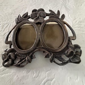 May include: A double oval picture frame crafted from dark, tarnished metal. The frame features an intricate relief design of pansy flowers and leaves surrounding two circular openings. The metal has a matte, aged finish with visible patina.