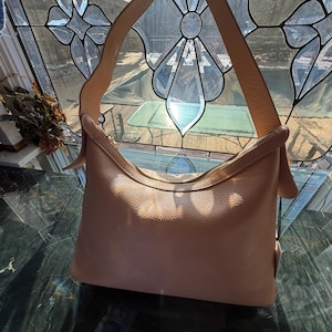 May include: A light brown leather shoulder bag with a top zipper closure and a single shoulder strap. The bag has a textured surface and is held up by a hand. The background features a stained glass window.