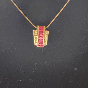 May include: A gold-toned necklace with a rectangular pendant. The pendant features a row of red gemstones and is accented with smaller clear stones. The necklace is displayed against a dark background.