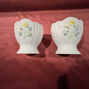 May include: Two white ceramic vases with a Celtic knot design and raised floral accents. The flowers are yellow, green, and blue.