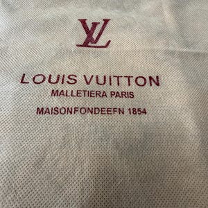 May include: Off-white fabric bag with a burgundy Louis Vuitton logo and text. The text reads "LOUIS VUITTON MALLETIERA PARIS MAISON FONDEE EN 1854". The bag has a textured surface.
