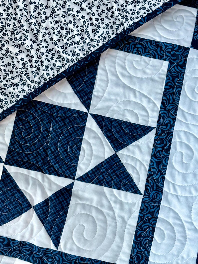 May include: Close-up of a handmade quilt with a patchwork design. The quilt features white and navy blue fabrics with floral and geometric patterns. The white sections have a spiral quilting pattern, and the edges are trimmed with a navy blue border.
