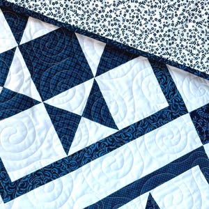 May include: Close-up of a quilted textile featuring a geometric star pattern in navy blue and white. The quilt has intricate stitching and a floral patterned border. The top corner displays a white background with a navy blue floral pattern.