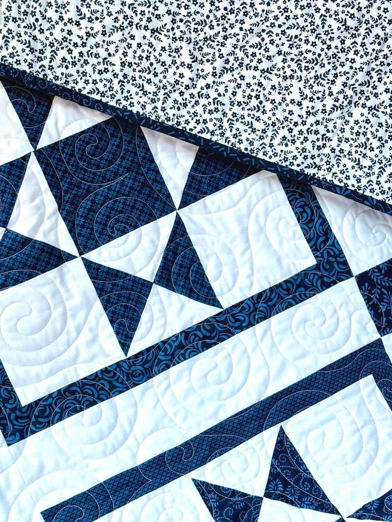 May include: Close-up of a quilted textile featuring a geometric star pattern in navy blue and white. The quilt has intricate stitching and a floral patterned border. The top corner displays a white background with a navy blue floral pattern.