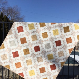 Pode incluir: Um edredom branco com um padrão geométrico de quadrados em tons de marrom, laranja e amarelo. O edredom é feito com um tecido estampado floral e tem uma textura acolchoada.